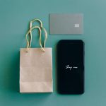 Vista superior de um smartphone com a mensagem “Shop now”, cartão de crédito e sacola de compras sobre fundo verde.