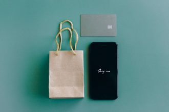 Vista superior de um smartphone com a mensagem “Shop now”, cartão de crédito e sacola de compras sobre fundo verde.