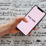 Mão segurando um smartphone com a mensagem “Stay Safe” sobre anotações manuscritas.