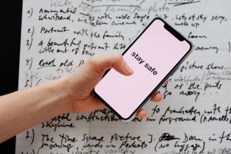 Mão segurando um smartphone com a mensagem “Stay Safe” sobre anotações manuscritas.